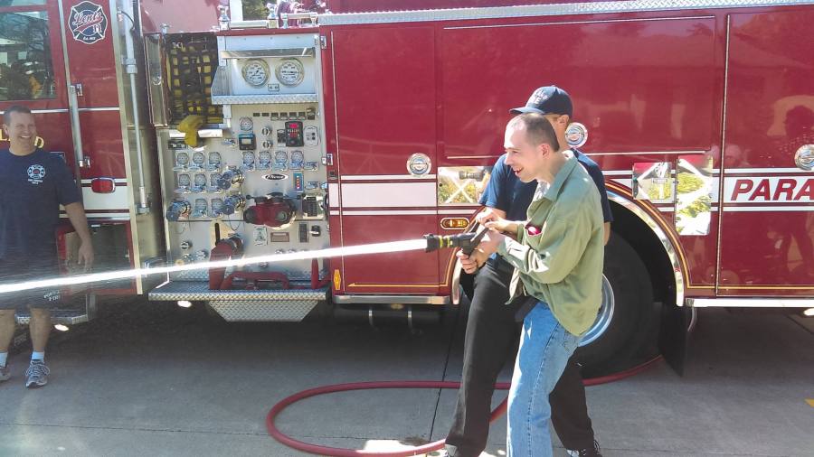 A client in front of a firetruck, spraying a fire hose while assisted by a firefighter.
