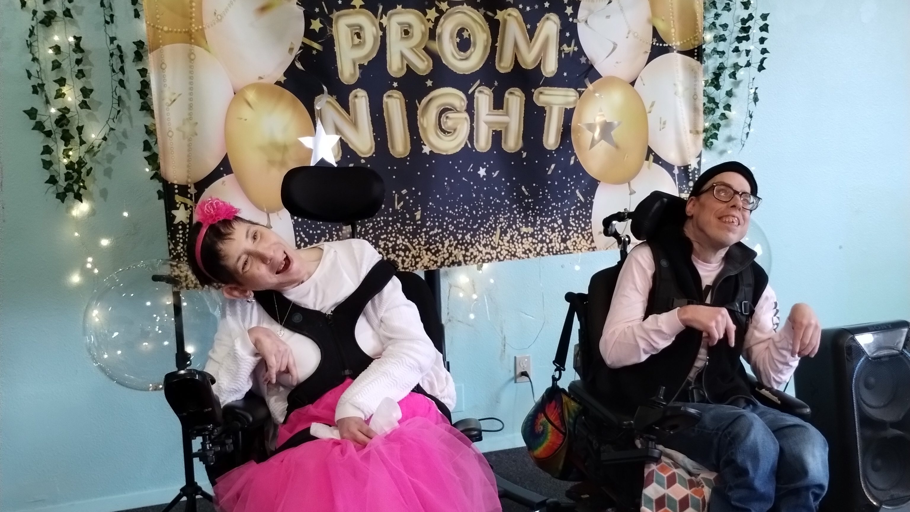 Two clients are smiling and seated in front of a banner with "PROM NIGHT" and balloons on it.